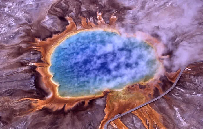 Aerial photograph of Grand Prismatic Spring in Yellowstone National Park, Wyoming, showing a vivid blue center surrounded by bright orange, golden, and purple mineral rings.