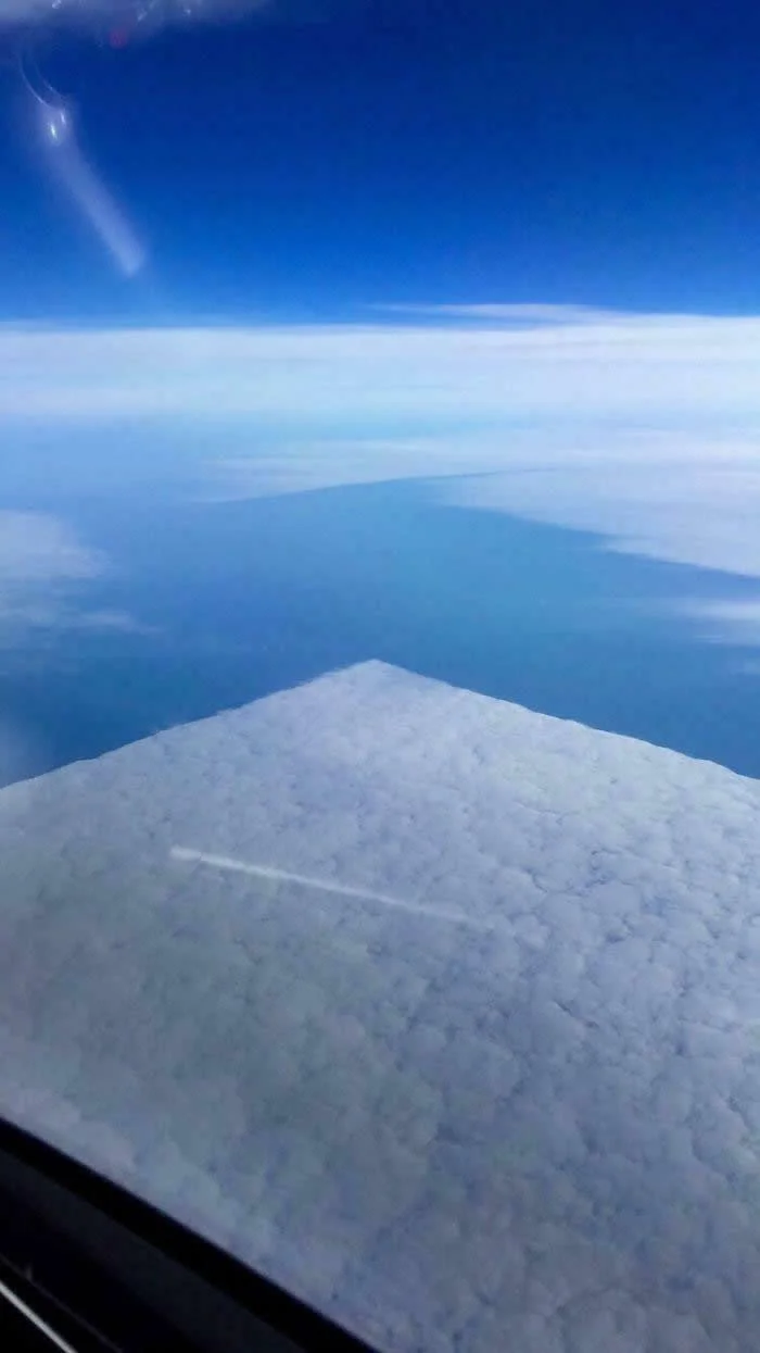 A large, square-shaped white cloud viewed from an airplane window, surrounded by layers of clouds and a deep blue sky above the Earth.