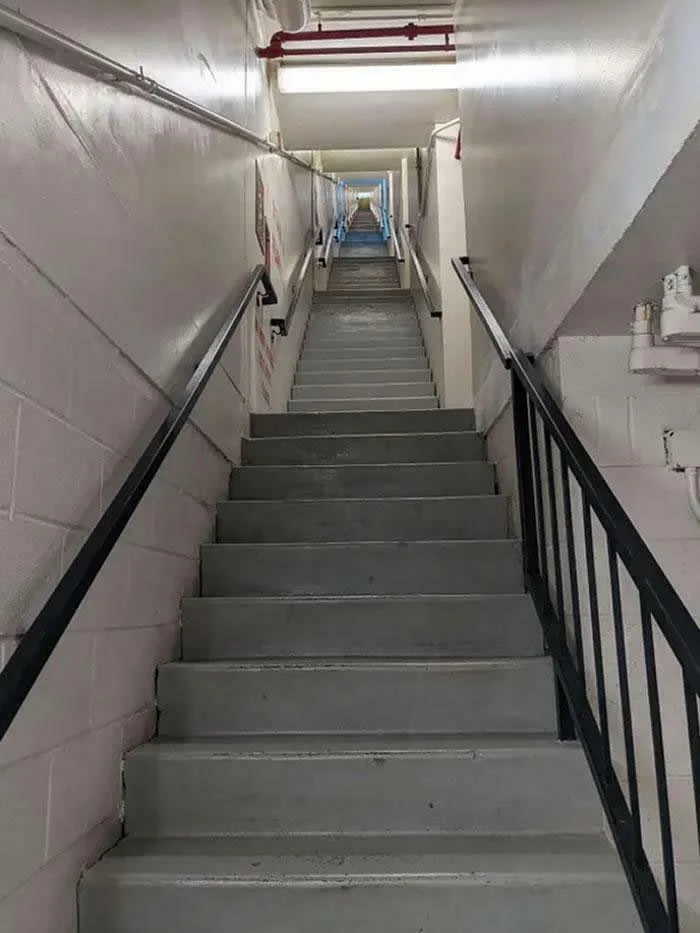 Long, straight concrete staircase inside a hotel, extending through multiple floors without switchbacks, with gray steps, white walls, black handrails, and exposed ceiling pipes.