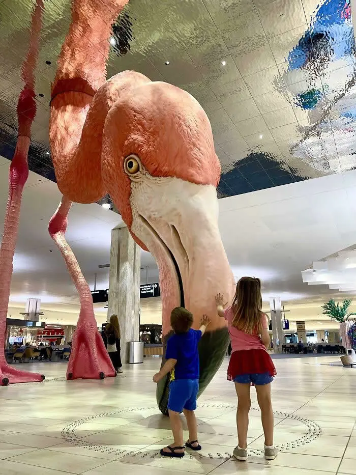 Giant pink flamingo sculpture at Tampa International Airport towers over two children touching its body beneath a reflective ceiling in the airport terminal.