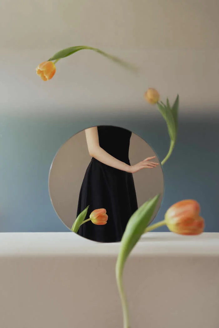 Surreal fine art photograph by Ziqian Liu showing a woman in a black dress reflected in a round mirror, surrounded by orange tulips against a soft beige and blue background.