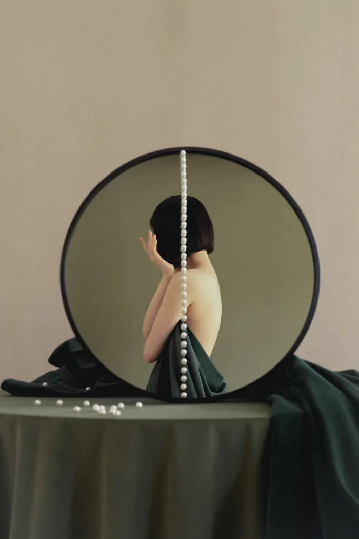 Surreal fine art photograph by Ziqian Liu showing a woman with short black hair in a dark green dress, reflected in a round mirror with a strand of white pearls running vertically through the composition.