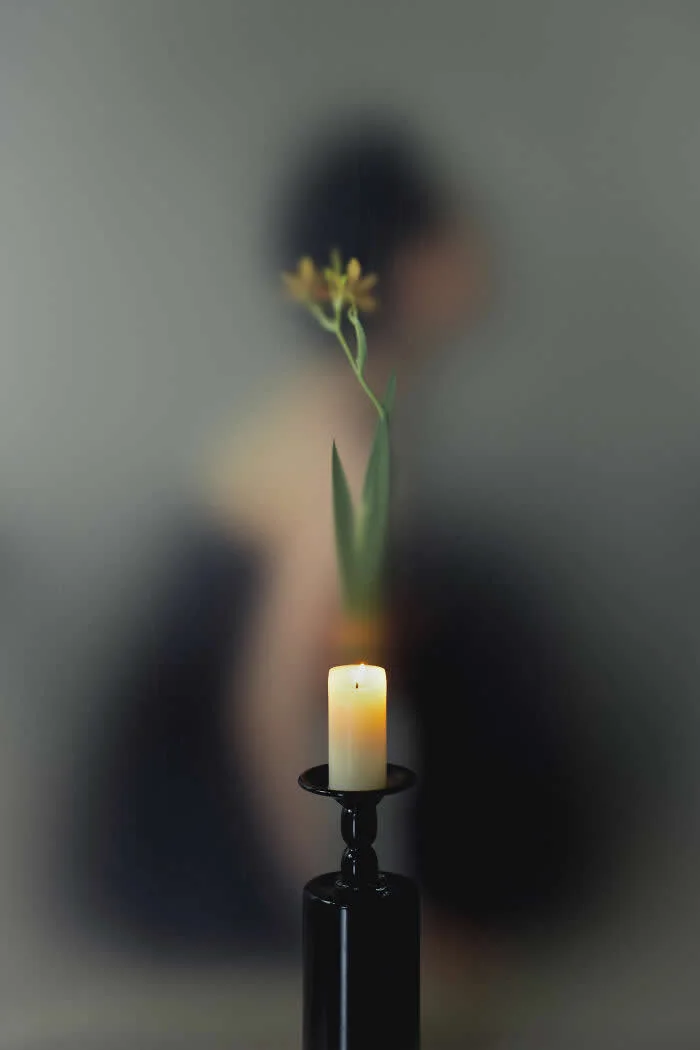 Surreal fine art photograph by Ziqian Liu featuring a lit candle on a black stand, a slender green plant with tiny yellow flowers, and a blurred human figure in the background.