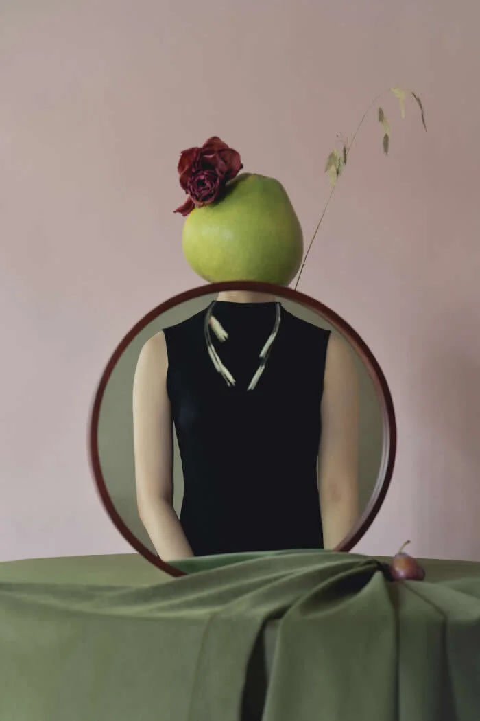 Surreal conceptual photograph by Ziqian Liu featuring a headless figure in a black sleeveless dress, reflected in a round mirror, with a green fruit topped by a dark red rose replacing the head. Green fabric covers the table.