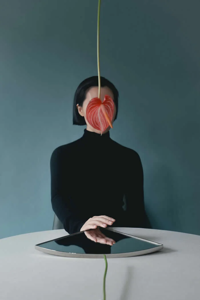 Surreal conceptual photograph by Ziqian Liu showing a woman in a black turtleneck seated at a white table, with a large red flower covering her face and a reflective tray beneath her hand against a muted blue background.