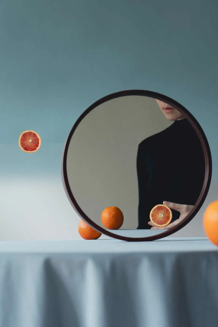 Surreal conceptual photograph by Ziqian Liu featuring a woman in black reflected in a round mirror, holding a halved orange, with whole oranges and another floating-looking orange slice arranged against a soft blue background.
