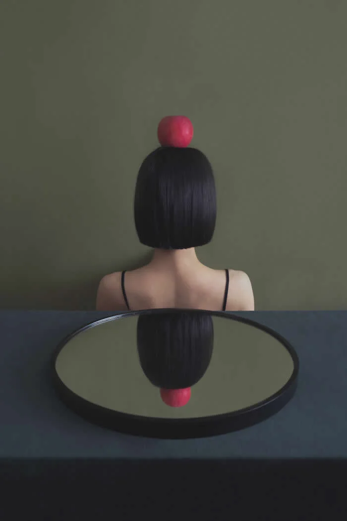 Surreal conceptual photograph by Ziqian Liu showing a woman with a short black bob haircut and a red apple balanced on her head, viewed from behind, with her reflection appearing upside down in a round mirror on a dark table.