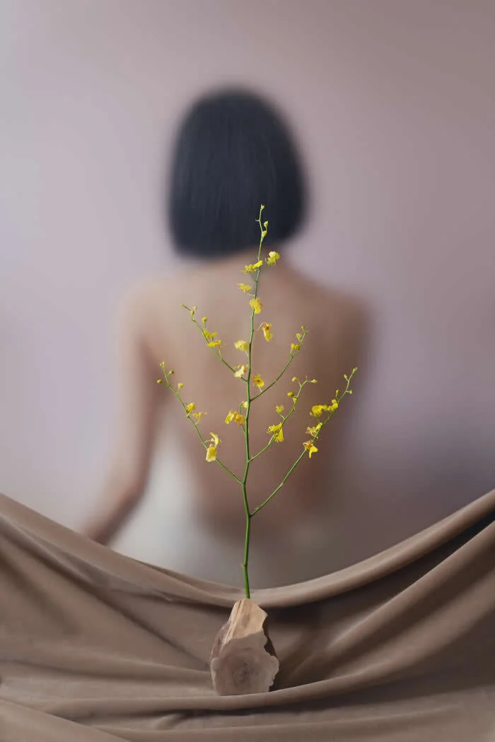 Surreal fine art photograph by Ziqian Liu showing delicate yellow flowers in sharp focus before a blurred woman with short black hair, surrounded by flowing beige fabric against a soft pink background.