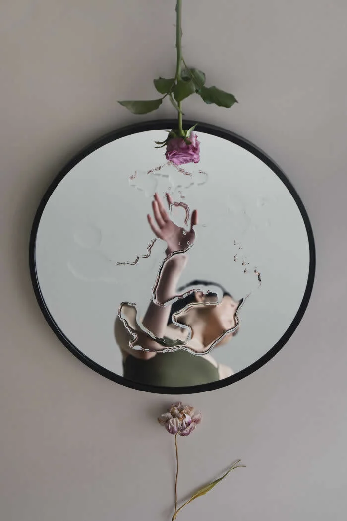 Surreal fine art photograph by Ziqian Liu showing a woman&rsquo;s distorted reflection reaching toward a pink rose above a circular mirror, with a delicate dried flower below against a minimalist beige background.