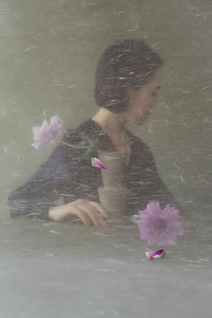 Surreal fine art photograph by Ziqian Liu showing a woman in a black top behind a textured translucent surface, surrounded by purple flowers and scattered petals against a muted gray-green background.