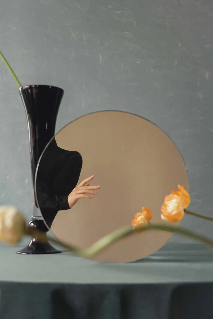 Surreal fine art photograph by Ziqian Liu featuring a round mirror, a tall black vase, orange flowers, and a partially obscured figure in black, arranged on a muted green table against a textured gray background.