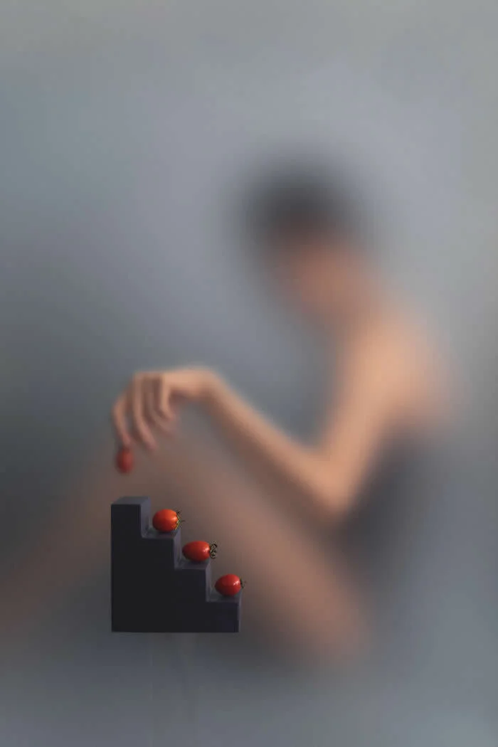 Surreal conceptual photograph by Ziqian Liu showing a blurred woman holding a small red object above three red tomatoes arranged on black stepped blocks against a soft blue-gray background.