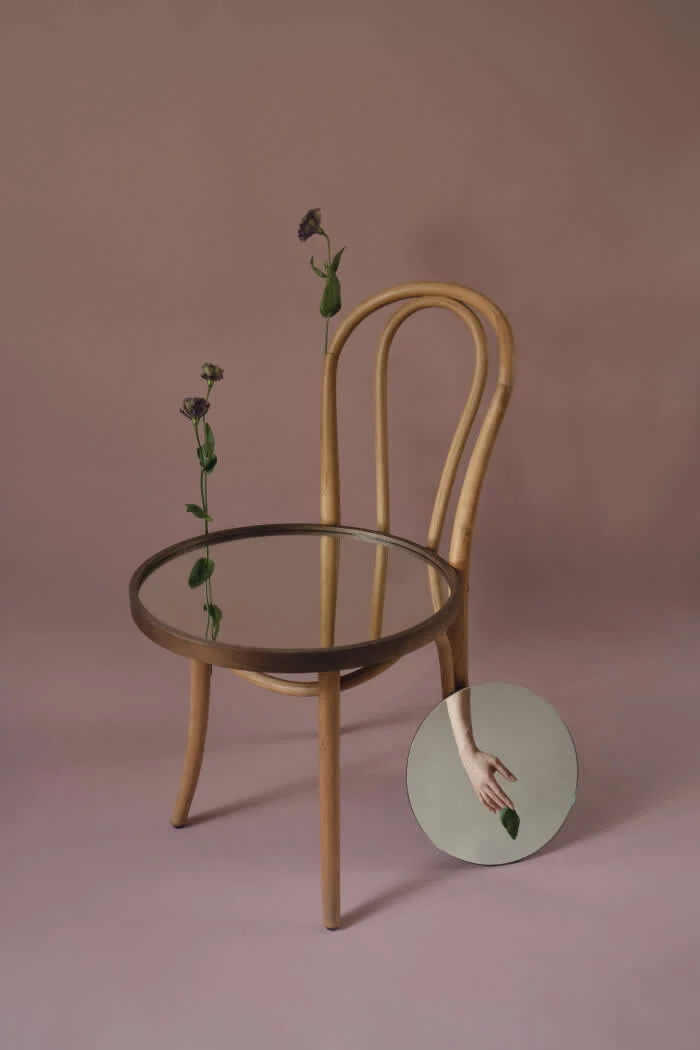 Surreal fine art photograph by Ziqian Liu featuring a wooden chair with a circular mirrored seat, two delicate flowers, and a small round mirror reflecting a hand holding a leaf against a muted pink background.