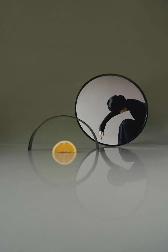 Surreal fine art photograph by Ziqian Liu showing a woman dressed in black bending inside a round mirror, with an orange slice beside an overlapping circular glass surface on a reflective table against an olive-green background.