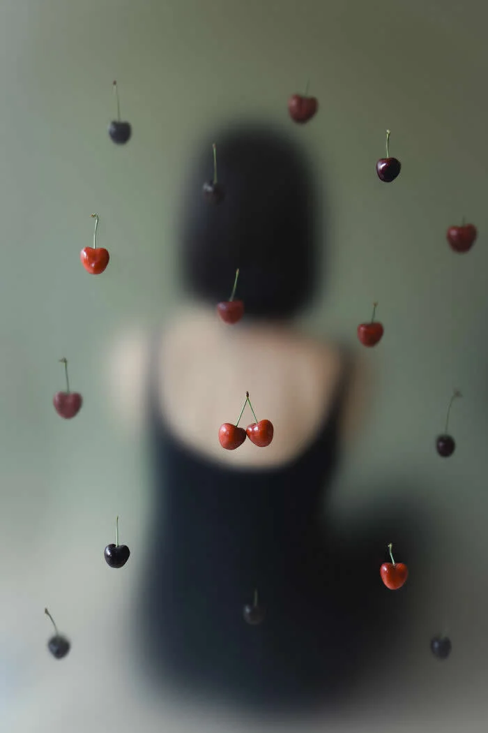 Surreal fine art photograph by Ziqian Liu showing red and dark cherries appearing to float in front of a blurred woman with short black hair, wearing a black dress against a soft green background.