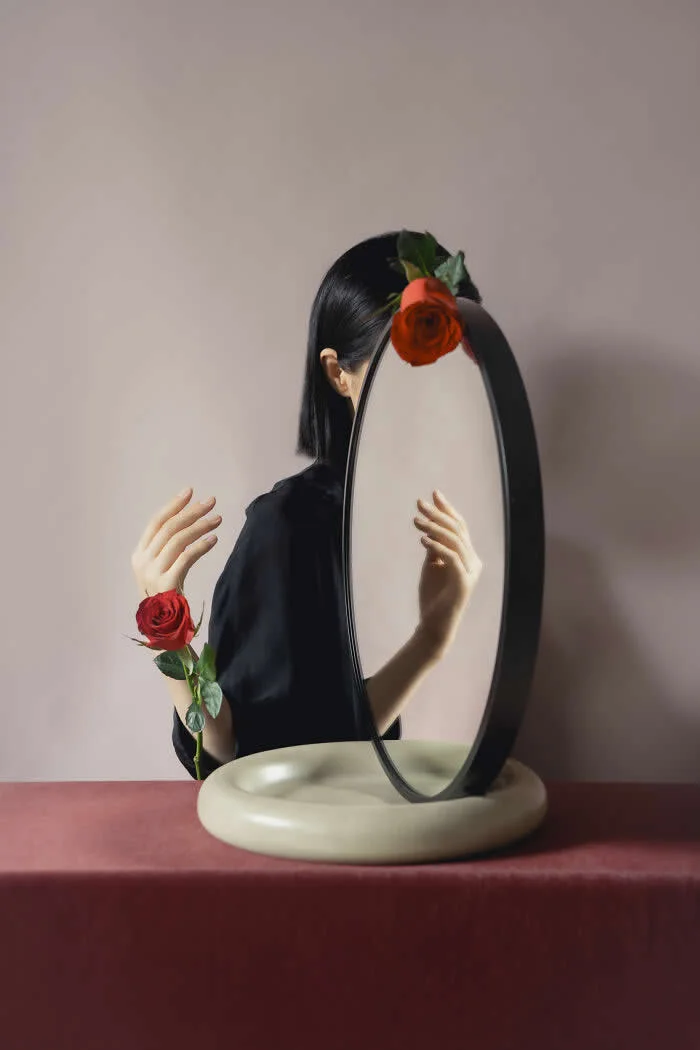 Surreal fine art photograph by Ziqian Liu showing a woman in a black outfit behind an oval mirror, with red roses positioned near her head and wrist, arranged on a cream-colored stand against a muted pink background.