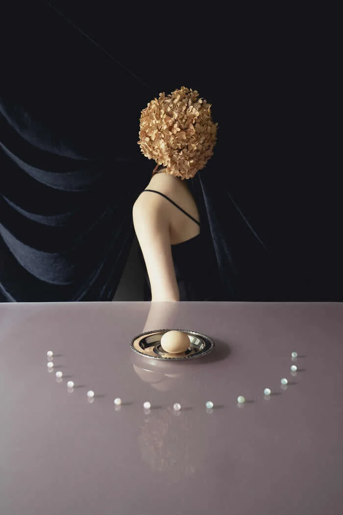 Surreal fine art photograph by Ziqian Liu showing a woman in a black dress with her face covered by dried flowers, standing behind a mauve table with an egg on a silver plate and small pearls arranged in a curved pattern.
