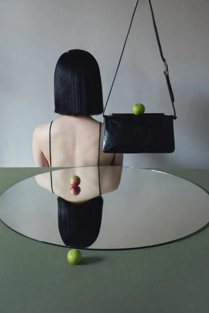 Surreal fine art photograph by Ziqian Liu showing a woman with a short black bob haircut viewed from behind, a black handbag holding a green apple, and a circular mirror reflecting her figure and colorful apples on a muted green table.
