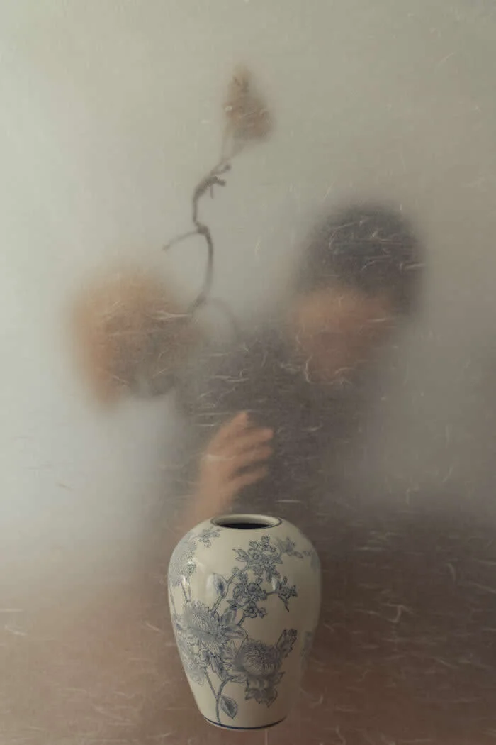 Surreal fine art photograph by Ziqian Liu showing a blue-and-white floral ceramic vase in the foreground, with a blurred woman and a delicate dried flower visible through a textured translucent surface against a muted beige background.