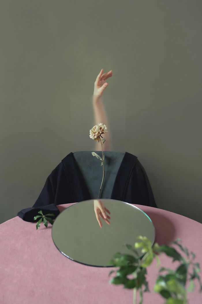 Surreal fine art photograph by Ziqian Liu showing a headless figure in black behind a pink table, with a dried flower rising from the neck, a raised hand above, and a circular mirror reflecting another hand.
