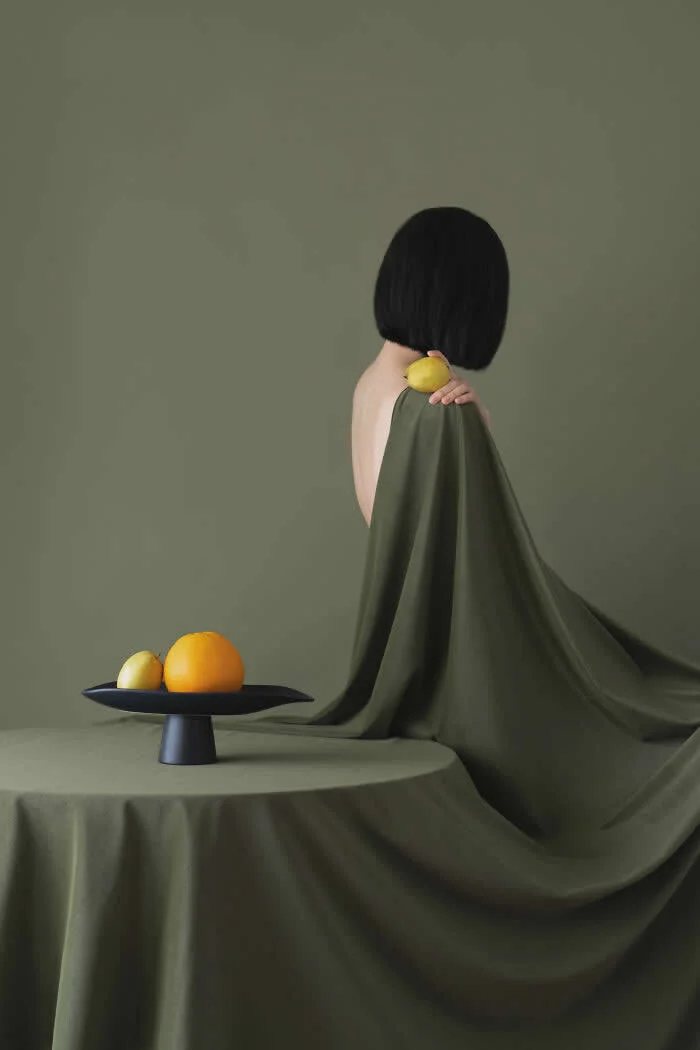 Surreal fine art photograph by Ziqian Liu showing a woman with a short black bob haircut draped in flowing olive-green fabric, holding a lemon near her shoulder, with oranges and lemons arranged on a black tray against a muted green background.