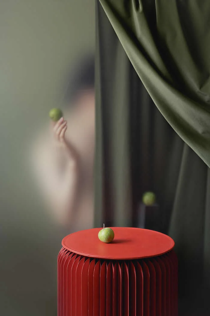 Surreal fine art photograph by Ziqian Liu showing green apples on a bright red sculptural table, with a blurred figure holding another apple behind translucent glass and flowing dark green curtains against a muted olive background.