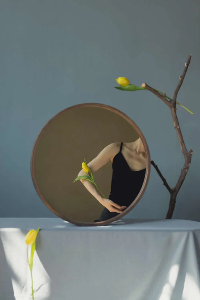 Surreal fine art photograph by Ziqian Liu showing a woman in a black dress partially reflected in a round mirror, surrounded by yellow tulips and a bare branch against a muted blue background.