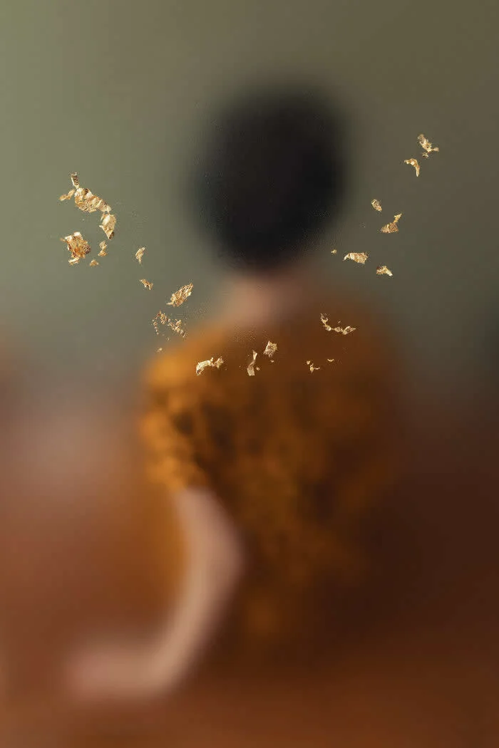 Surreal fine art photograph by Ziqian Liu showing a blurred figure in warm brown clothing against an olive-green background, surrounded by scattered golden fragments floating across the foreground.