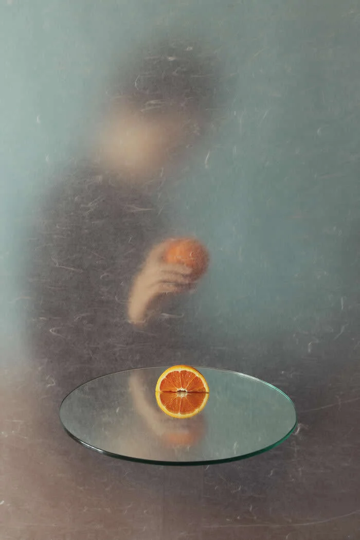 Surreal fine art photograph by Ziqian Liu showing a blurred woman holding an orange behind a textured translucent surface, with a sliced orange resting on a round glass platform against a muted blue-green background.