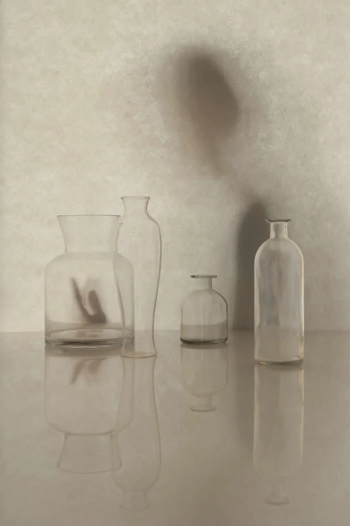Surreal fine art photograph by Ziqian Liu showing four transparent glass bottles reflected on a glossy surface, with a blurred hand-shaped shadow visible through one bottle against a textured beige background.