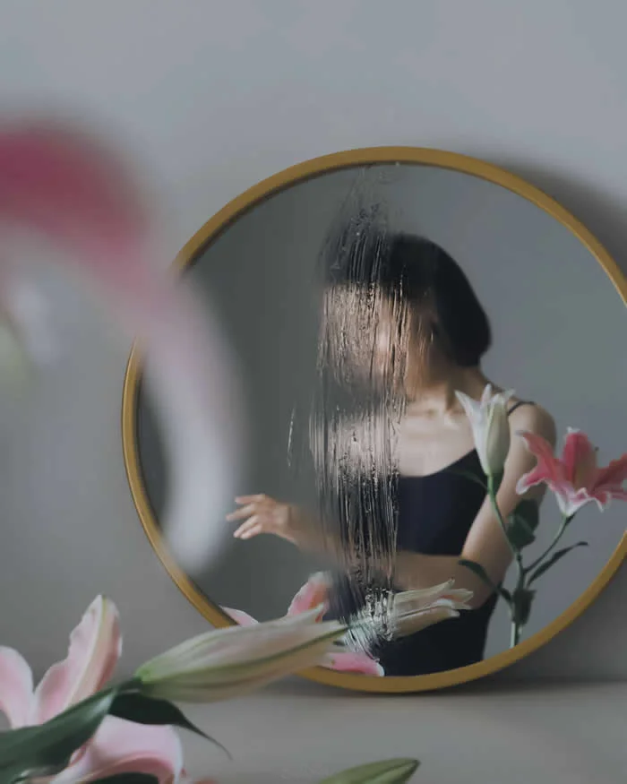 Surreal fine art photograph by Ziqian Liu showing a woman in a black top reflected in a round gold-framed mirror, partially obscured by textured glass and surrounded by pink lilies against a soft gray background.
