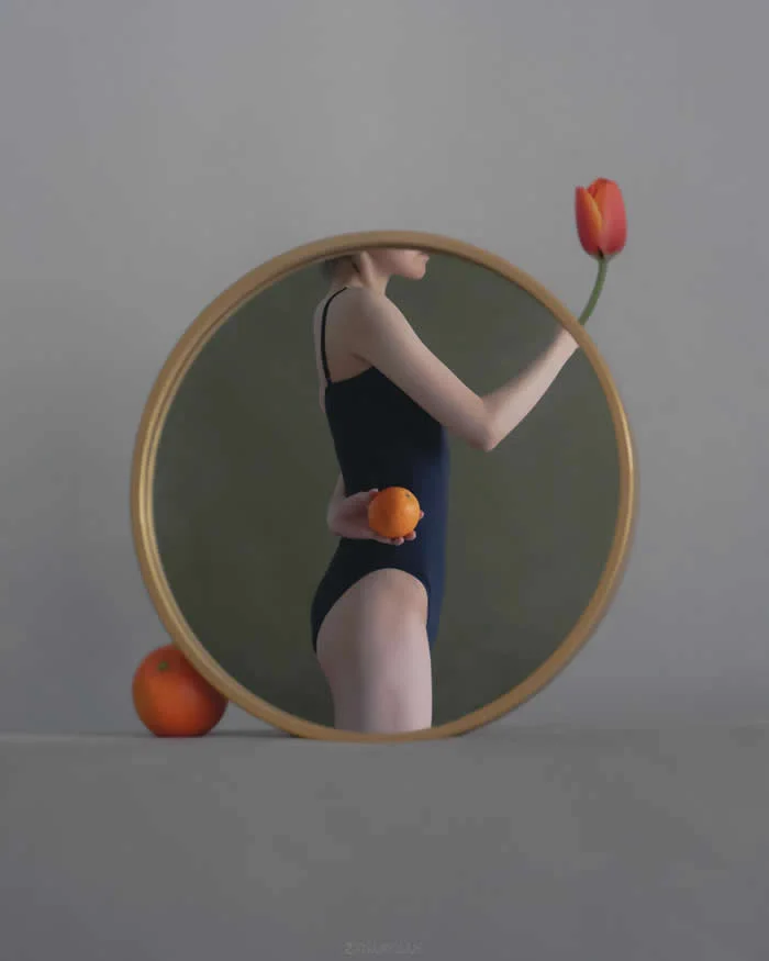 Surreal fine art photograph by Ziqian Liu showing a woman in a black swimsuit reflected in a round gold-framed mirror, holding an orange behind her back and reaching toward a red tulip, with another orange beside the mirror against a gray background.