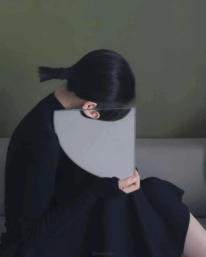 Surreal fine art photograph by Ziqian Liu showing a woman in a black dress sitting with her head bowed, holding a curved rectangular mirror that obscures part of her face, against a muted olive-green and gray background.