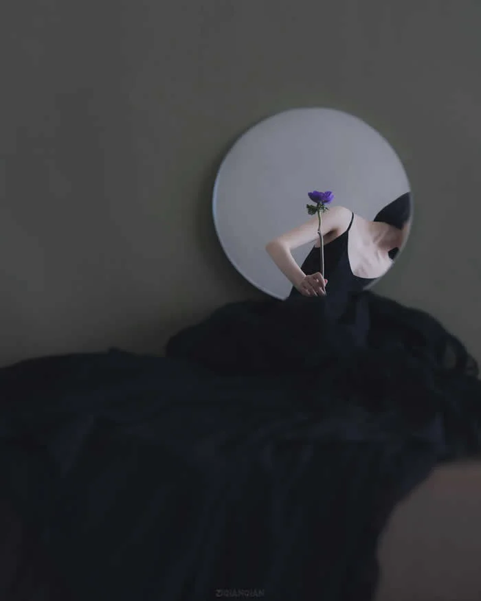 Surreal fine art photograph by Ziqian Liu showing a woman in a black dress holding a small purple flower inside a round mirror, with her reflection partially visible against a dark gray background and flowing black fabric.