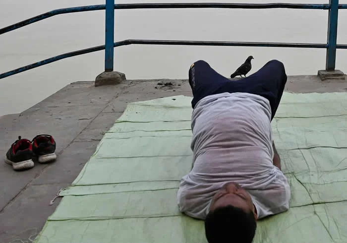 Perfectly Timed Animal Photos: 40 Funny and Unexpected Moments by Pranay Pariyar 52 A man lies on a green mat near a waterfront with his knees bent, while a black crow perches between his legs and a pair of shoes rests nearby.