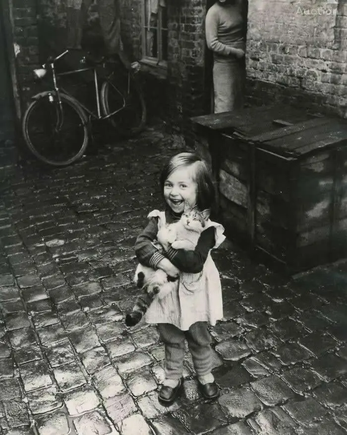 Smiling young girl holding a cat in a wet cobblestone alley, with a bicycle, brick walls, and adults in the background, captured in a vintage black-and-white documentary photograph
