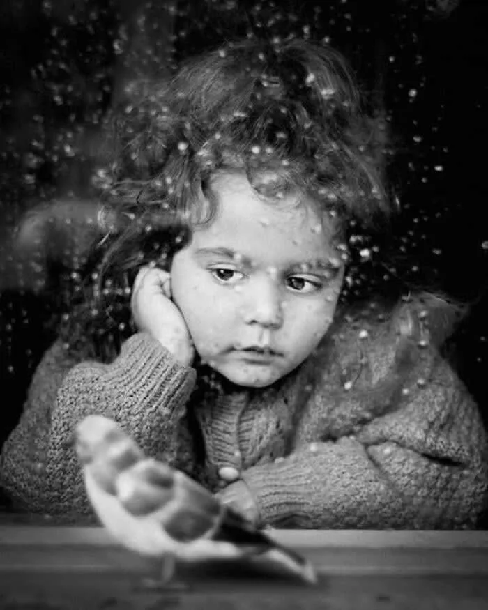 Black-and-white portrait of a young child watching a bird through a rain-speckled window, with the child&rsquo;s face in focus and the bird softly blurred in the foreground.