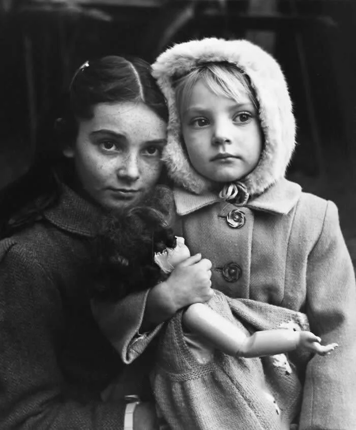 Black-and-white vintage portrait of two children standing closely together, with one holding a doll and the younger child wearing a fur-trimmed coat and looking toward the camera.