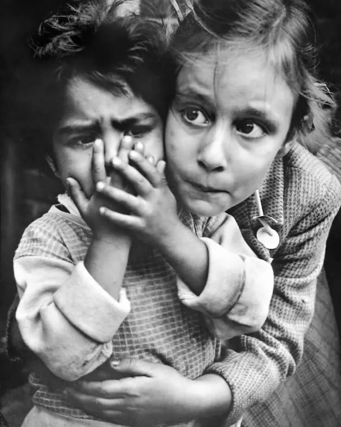 Black-and-white vintage photograph of two children embracing, with one child covering the other&rsquo;s mouth while both look toward the camera.