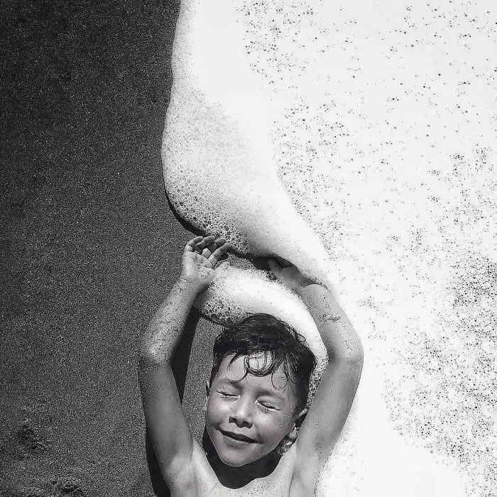 Black-and-white overhead photograph of a smiling child with raised arms beneath a large foamy wave, creating an abstract composition of water, movement, and childhood joy.