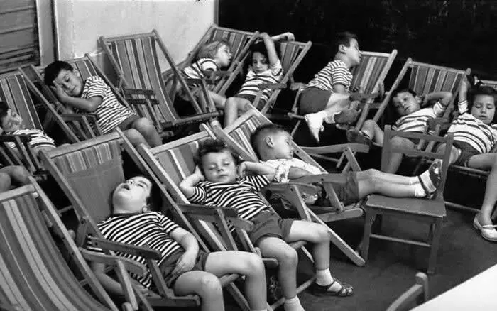 Group of children relaxing and lounging in striped deck chairs, with several appearing sleepy or resting, captured in a playful vintage black-and-white photograph.