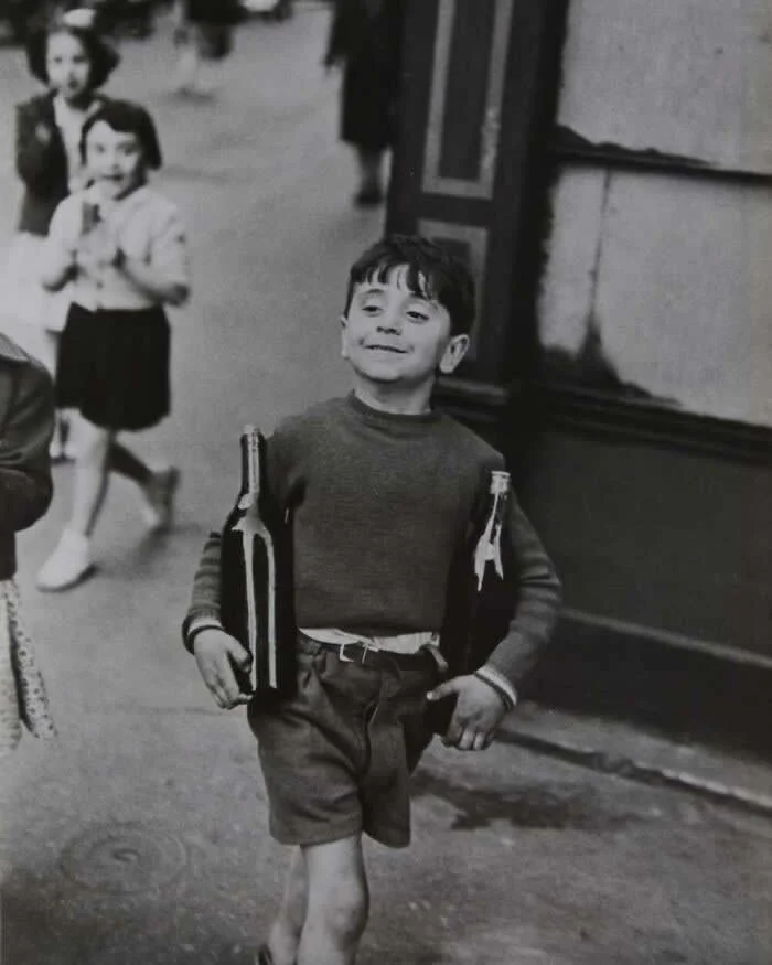 Cheerful young boy walking down a street while carrying two large glass bottles under his arms, with other children visible in the background, captured in a vintage black-and-white photograph.