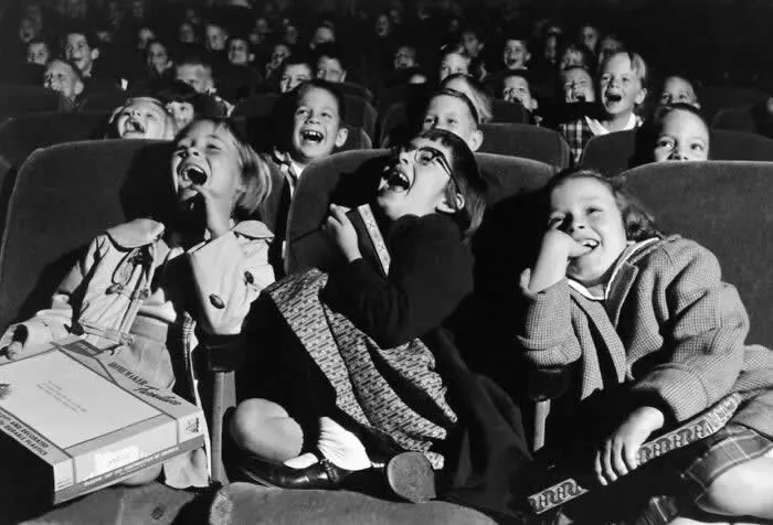 Group of children laughing and smiling together in rows of theater seats, captured during a lively moment in a vintage black-and-white photograph.