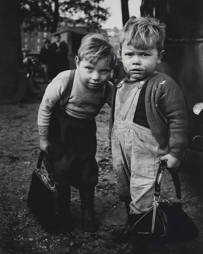 Two young boys standing together with school satchels in a muddy outdoor setting, wearing vintage clothing and looking seriously toward the camera in a black-and-white documentary photograph.