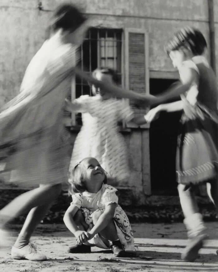 Three children playing outdoors, with two girls moving around a younger child sitting on the ground, captured with expressive motion blur in a vintage black-and-white photograph.