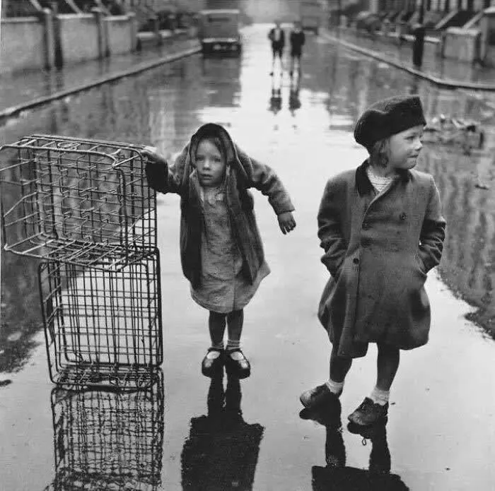 Two children walking along a rainy city street beside an empty shopping cart, with their reflections visible in puddles and distant pedestrians in the background, captured in black and white.