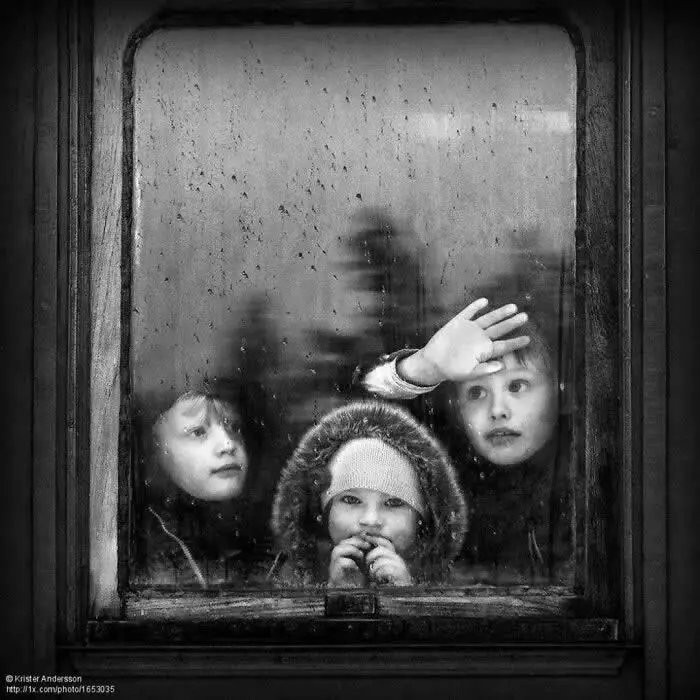 Three children looking through a fogged, rain-speckled window, with one child raising a hand and another resting their fingers near their mouth, captured in a moody black-and-white photograph.