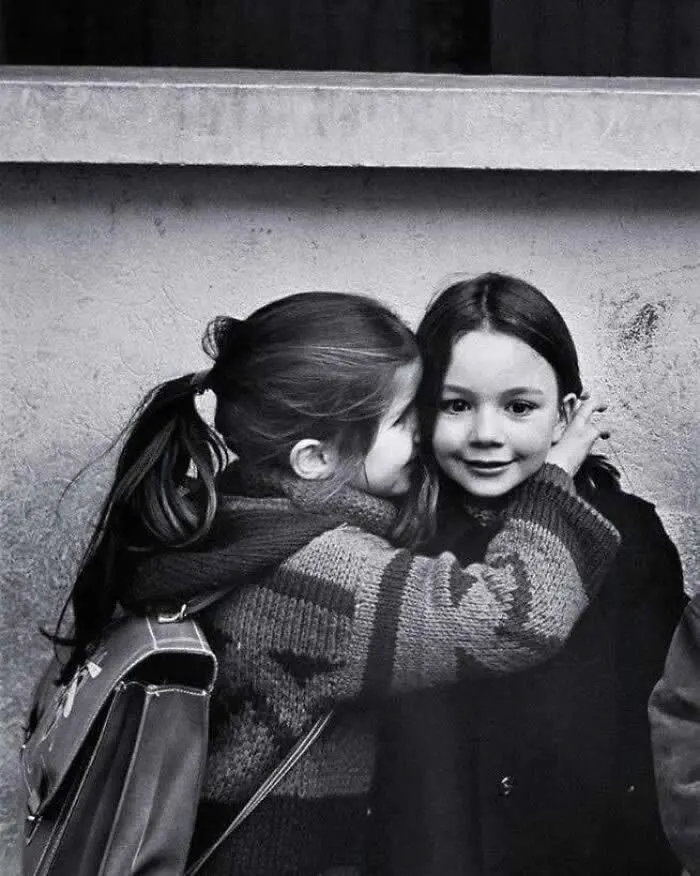 Two young girls embracing, with one affectionately leaning toward the other&rsquo;s cheek, photographed against a simple wall in a vintage black-and-white portrait.