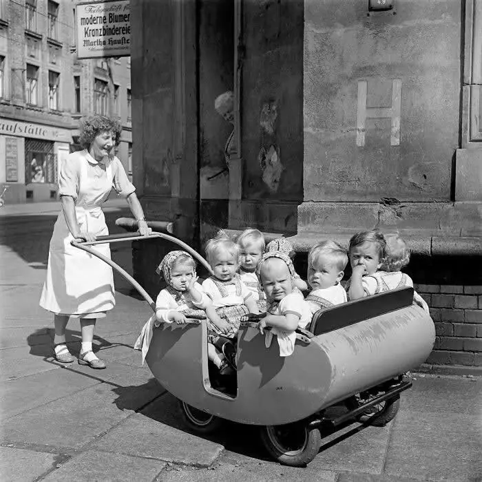 Woman pushing a large vintage pram filled with several young children along a city sidewalk, surrounded by historic buildings and street signs in a black-and-white photograph.