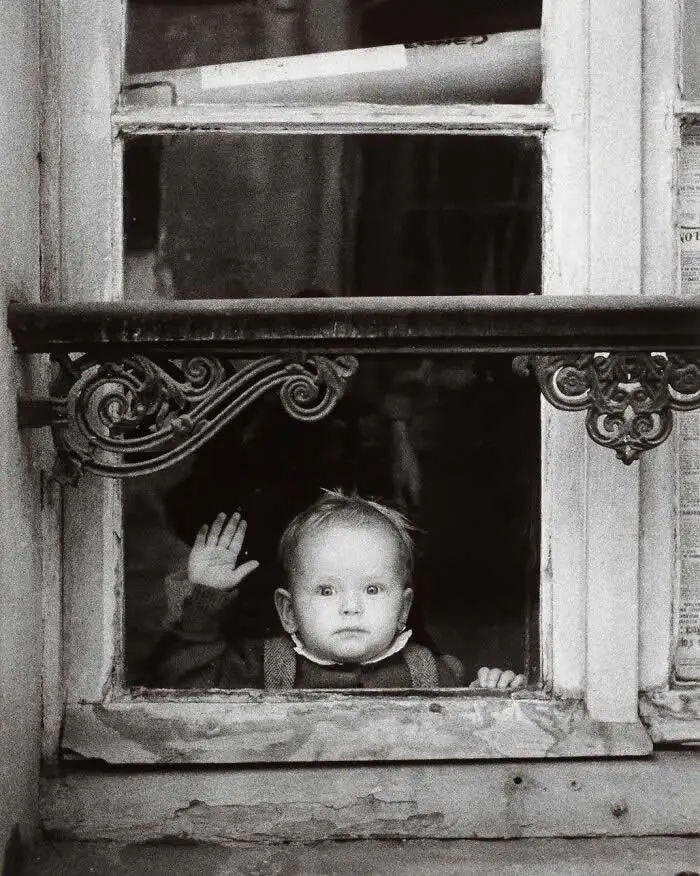 Baby looking through an old window and raising one hand, framed by ornate ironwork and weathered wood, captured in a vintage black-and-white photograph.